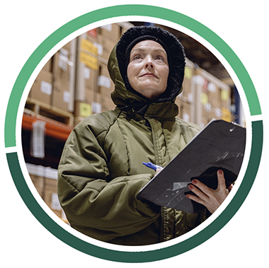 light skinned person in a green coat writing on a clipboard in a warehouse